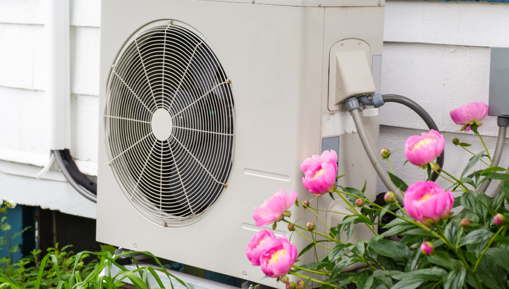 A heat pump outdoor unit installed next to a home with white siding. Pink flowering bush in foreground.